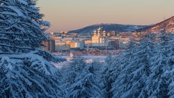 Amazing winter landscape. View from the snow-covered forest to the northern city and mountains. Snow on the branches of larch trees. Beautiful cathedral at sunrise. Cold snowy weather. Magadan, Russia