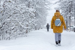 People walk on a snow-covered sidewalk during a heavy snowfall. Lots of snow on the ground and branches of trees and bushes. Cold snowy winter weather. Woman in warm winter clothes with a backpack.