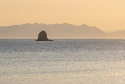 Morning seascape. View of the sea bay and a rock in the sea. Mountains in the distance. Golden sunlight at sunrise. Gertner Bay, Sea of Okhotsk, Magadan Region, Far East of Russia. Natural background.