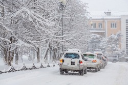Cars drive along a snow-covered city street. Heavy snowfall and snowstorm in the city. A lot of snow on the roadway, cars and trees. Cold snowy winter weather. Magadan, Siberia, Russia.