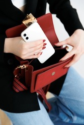 girl puts white smartphone in small purse. small red purse. Close-up of a young girl using a cell phone by putting a cell phone in her purse. 