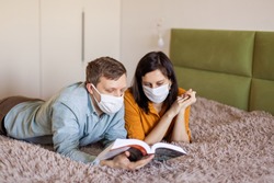 A young couple is sitting at home on self-quarantine. Husband and wife quarantined  coronavirus in protective masks. New reality. Normal life in isolation. Read a book together. Italy europe covid-19