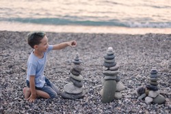 Happy boy builds a cairn of stones on the sea coast. Beach games with children. Leisure in the evening on vacation with kids. Balance development building fantasy. Childhood. Fun joy. Ocean trawel