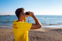 Lifeguard on the beach looking through binoculars. Safety while swimming, handsome brunette male lifeguard on the beach,back shot looking at the blue sea, copy space.