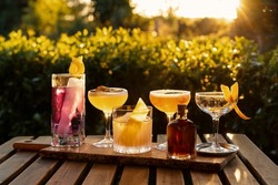 Selection of five gourmet cocktails in different fancy glasses with dried orange peel and colourful liqueurs on a wooden board and wooden table with sunset light and a bottle of red spirit