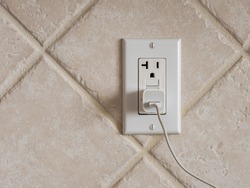 USB cell phone power adapter brick and cord in GFCI wall plug. Electrical cord and charger plug in wall outlet.