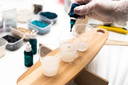 Epoxy resin. A tube of blue dye in the artist's hand, against the background of three glasses with resin, standing on a wooden board