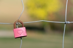 A small padlock hangs on a wire fence. The padlock is closed. The color is pink. A heart is engraved on the lock. The metal bracket is rusty. In the background washed out green landscape.