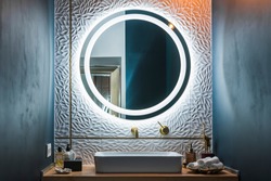 Modern bathroom interior with white wash basin, golden faucet and round illuminated mirror.