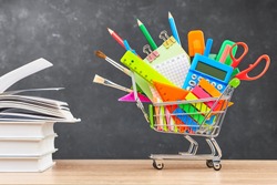 Various office supplies for school in a cart and textbooks on a wooden table on a blackboard background. The concept of preparing for school, the beginning of a new school year.