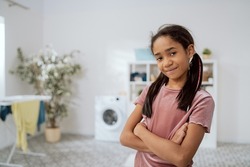A little girl with dark skin and hair stands in tshirt in middle of laundry room, the bathroom with arms crossed over her chest and a smile, in the background a washing machine, clothes dryer