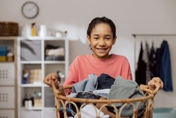 Smiling pretty girl stands in the middle of the bathroom, laundry room, holding a large wicker basket filled with colorful clothes in hands, daughter helps mother with household chores.