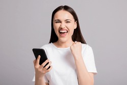 Portrait of young attractive adorable lovely pretty cute successful cheerful woman winning celebrating isolated on grey background holding cellphone showing delighted emotions at camera.