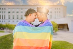 Couple of gay men in love embracing each other, wrapped in a gay flag, in a park at sunset, with reflections of the sun. Concept of diversity, pride, love, equality.
