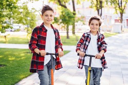 Two attractive european boys brothers, wearing red and white checkered flannek shirts, standing on scooters in the park. They laughing, smiling, hugging and having fun. Showing big fingers up sign.