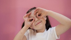 Young dark hair girl with funny ponytails makes glasses from fingers and smiles, looks at the camera. Pink background