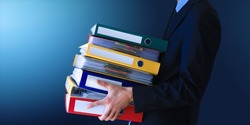 Professional business man holding heavy huge stack of ring binders for archiving documents over blue isolated background. Heavy folder full of paper documents. 