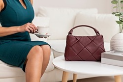A beautiful young woman poses in a green dress against the background of a sofa in the living room. A woman is holding a burgundy shoulder bag made of braided leather. The concept of women's fashion.