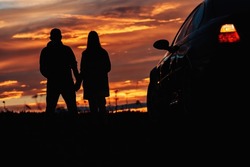 A silhouette of a couple standing near a car against the backdrop of a beautiful sunset or dawn sky.