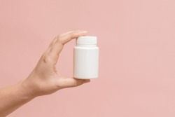 White bottle (plastic tube) in hand on a pink background. Packaging for vitamins, pill or capsule, or supplement. Mockup for product branding