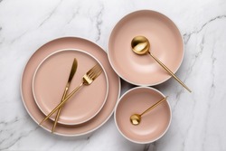 Dishes and utensils for serving and eating meals. Beige round rimmed plates and gold colored cutlery on a white marble table, top view. Modern ceramic crockery, trendy tableware