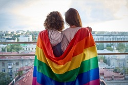 Two girls with rainbow flag looking out the window at the city. Lesbian couple celebrating pride month during Covid-19 quarantine, stay at home
