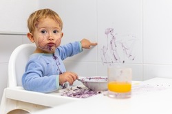 blond boy in his high chair shows the wall that he has stained with his breakfast