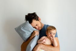 Young father holding his baby in one hand and a pillow in other hand looking tired, trying to sleep.