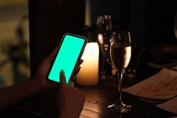 close up people tapping green screen smart phone in the bar at night. blurred glass of wine and candle light as background