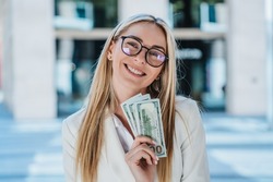 Excited blonde caucasian woman in glasses, white suite holding US dollar banknotes smiling wide, satisfied by profit against blurry buildings. Happy entrepreneur celebrates great deal, received cash.
