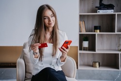 Dreamy blonde young Swedish woman holds credit card to friend, wants to order products via internet. Successful businesswoman at home making online purchase, holding phone. Shopping and online payment