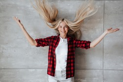 Beautiful  caucasian blonde woman in plaid shirt throwing her long hair in the air against marble wall, satisfied by her health. Body care concept. Cheerful model at studio. Pretty housewife at home.