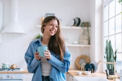 Happy caucasian female with long loose hair in jeans and blue shirt toothy smiles holds cup of tea looks aside happily enjoys weekend at cozy home. Pretty hispanic woman laughs against blurry kitchen.