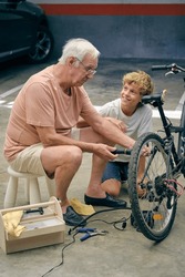Concentrated old grandfather screwing bolt while repairing bicycle with pleasant boy sitting close on parking lot