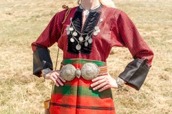 Old traditional women's clothing. A young girl - a woman in Bulgarian folk costume. Silver ornaments, red robe and silver belt. Women's clothing up close. Bulgarian folklore. Female hands. Sunny day
