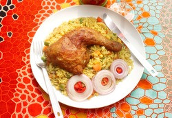  A Meal of Nigerian Fried Rice served with fried Chicken thighs and garnished with onion rings and chopped peppers. In a white plate on a colorful pattern background