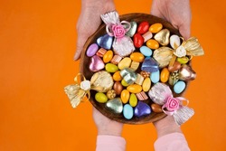 Holding bowl of sweet, woman and child hands holding bowl of sweet. Top view image of wrapped luxury chocolate and almond dragee in wooden tray. Isolated orange studio background with copy space.