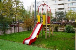 A wooden slide of bright red and yellow color against the background of green trees and residential buildings on a clear sunny day. Playgrounds, sports, entertainment.