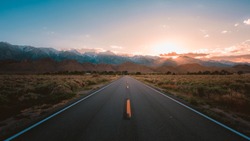 A straight road in the middle of the desert with magnificent mountains and the sunset in the background