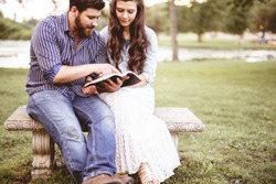 A closeup shot of a couple sitting on a bench and reading the bible with a blurred background