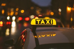 Illuminated taxi sign on top of a car. City lights with neon color in the background. Rainy urban night scene.
