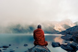 A Tibetan monk from back sitting on the stone near the water in the background of foggy mountains