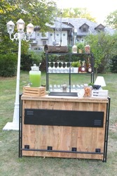 A vertical shot of a wooden bar full of glasses and drinks next to a lantern in green garden with background of a house