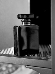 A grayscale closeup shot of a black perfume bottle on a metal mesh shelf with blurred background