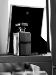 A grayscale closeup shot of a black perfume bottle on a metal mesh shelf with picture frame in the blurred background