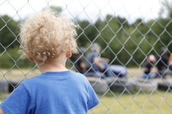 A backshot of a baby boy 3 years old standing behind a grid fence in daylight looking at a race track