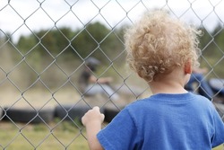 A backshot of a baby boy 3 years old standing behind a grid fence in daylight looking at a race track