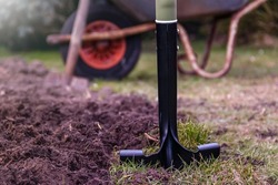 A closeup shot of a spade stuck in soil on the background of a wheelbarrow and trees