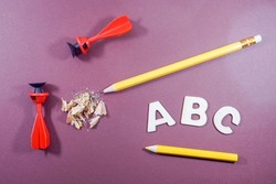 A top view of ABC letters in white, pencil, sharpener residue, yellow color pencil and two sucker darts on a purple background