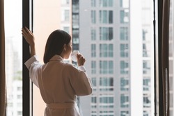 A Thai woman wearing a bath robe drinking coffee and admiring the scenery in a hotel room in morning
